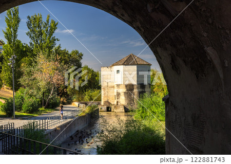 Cordoba town in Spain. Scenic view of a historic stone tower beside a calm waterway, seen through an archway. Cordoba town in Spain. Scenic view of a historic stone tower beside a calm waterway, seen through an archway. 122881743
