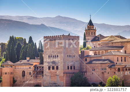 View of the Alhambra palace from the Mirador de San Nicolas lookout in Granada town in Spain. Ancient fortress nestled in mountains under a clear blue sky. 122881757