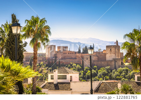 The Alhambra palace from viewpoint in Granada town in Spain. Historic castle nestled in a sunny, palm-tree filled landscape, mountains in the background. 122881765