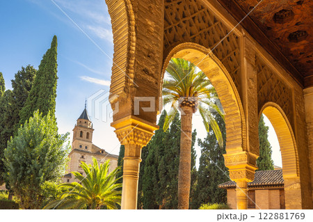 Alhambra complex in Granada town, Andalusia, Spain. Ornate arches frame a sun-drenched courtyard, revealing palm trees and a distant tower. 122881769