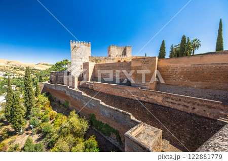 The Alcazaba fortress at the Alhambra complex in Granada town, Andalusia, Spain. Ancient fortress walls and towers against a clear blue sky. Lush greenery and distant hills complete the scene. 122881779
