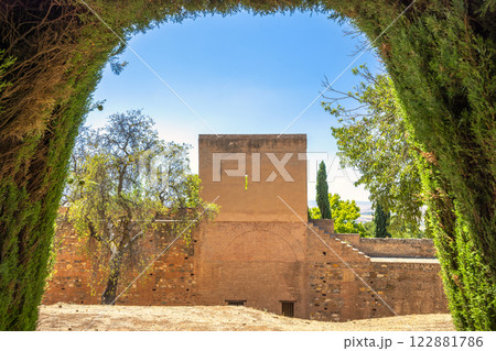 Alhambra complex in Granada town, Andalusia, Spain. Ancient stone wall viewed through a lush green archway, under a clear blue sky. A tranquil, historical scene. 122881786