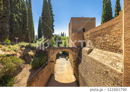 Aqueduct of the Acequia Real in the Alhambra complex, Granada town, Andalusia, Spain. Ancient stone archway and fortress walls under a clear blue sky. Tall cypress trees frame the scene. Aqueduct of the Acequia Real in the Alhambra complex, Granada town, Andalusia, Spain. Ancient stone archway and fortress walls under a clear blue sky. Tall cypress trees frame the scene. 122881790