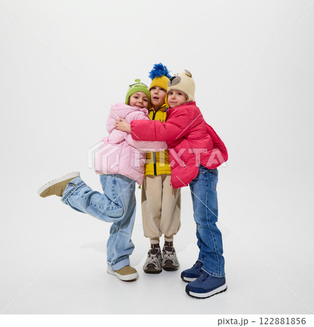 Three children, dressed warm winter jackets, hugging each other with smiles, showing friendship and joy of childhood against white studio background. 122881856