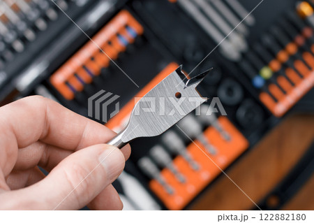 Hand holding a spade drill bit over an orange toolset. 122882180
