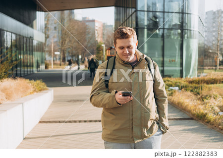 Handsome young man using phone outdoor. Smiling businessman walking outdoors using mobile in urban area. Urban lifestyle concept. Spring time. Handsome young man using phone outdoor. Smiling businessman walking outdoors using mobile in urban area. Urban lifestyle concept. Spring time. 122882383