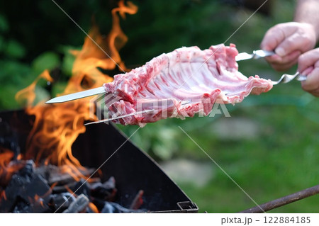 Pork ribs are skewered and positioned over a vibrant fire in a backyard. The intense flames create a warm and inviting atmosphere, perfect for a summer barbecue gathering with friends. Pork ribs are skewered and positioned over a vibrant fire in a backyard. The intense flames create a warm and inviting atmosphere, perfect for a summer barbecue gathering with friends. 122884185