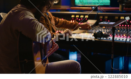 Creative musician singing guitar in professional studio control room, recording his tracks with producer. Singer songwriter composing a new song for his album, editing tunes on console. Camera A. Creative musician singing guitar in professional studio control room, recording his tracks with producer. Singer songwriter composing a new song for his album, editing tunes on console. Camera A. 122885010