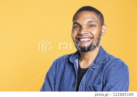Portrait of charming cool african american man posing on camera, standing with arms crossed against yellow background. Relaxed confident person in a good mood smiling in studio. 122885100