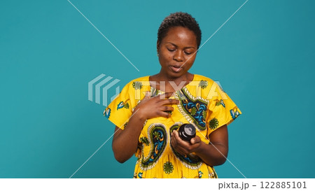 Young adult in pain dealing with a headache and looking for painkillers, reading a tag on a pills bottle against blue background. Sick girl takes pharmaceutical treatment for an illness. Camera A. 122885101