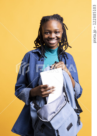 Portrait of upbeat BIPOC young girl browsing through backpack, isolated over studio background. Radiant college student taking out textbooks out of rucksack used during commuting to university 122885103