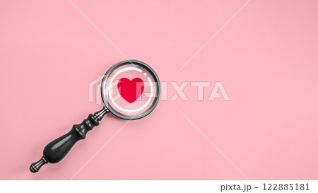 Black magnifying glass with a red heart icon inside, placed against a pastel pink background, symbolizing love in Valentine, romance, and the concept of searching for single people connection. 122885181