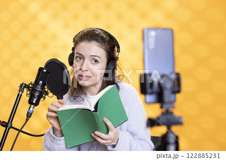 Influencer looking at phone camera, filming herself reading book, creating audiobook, isolated over studio background. Content creator doing vlog while producing digital recording on novel 122885231