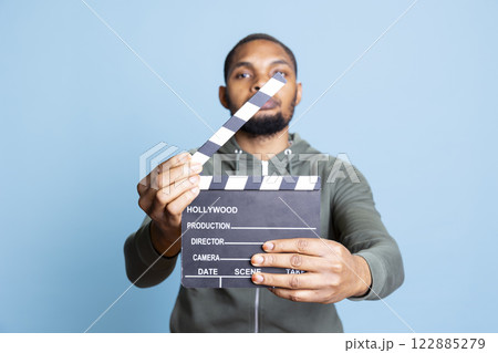 Smiling african american film producer posing with a clapperboard, cinematography industry tool from the set. Using the movie slate to cut scenes and start new takes for a cinema masterpiece. Smiling african american film producer posing with a clapperboard, cinematography industry tool from the set. Using the movie slate to cut scenes and start new takes for a cinema masterpiece. 122885279