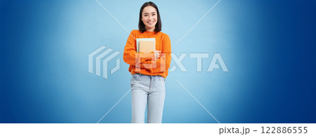Education and students. Happy asian woman, holding notebooks and laughing, smiling at camera, enjoys going to University or College, blue background Education and students. Happy asian woman, holding notebooks and laughing, smiling at camera, enjoys going to University or College, blue background 122886555