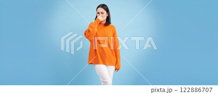 Something stinks. Young asian woman shuts her nose with fingers from bad smell, wears orange sweatshirt, stands over blue background 122886707