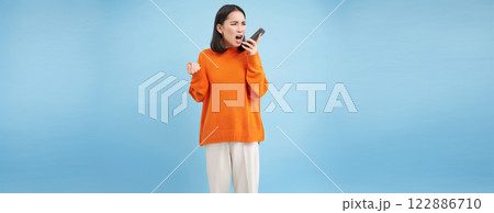 Angry asian woman yells at mobile phone, shouts in smartphone with furious face, stands over blue background 122886710