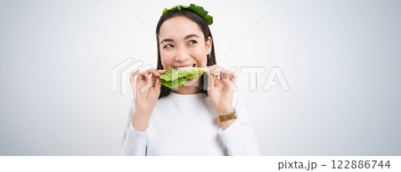 Close up portrait of smiling asian woman eating lettuce, loves cabbage, vegetarian enjoys raw organic food, white background Close up portrait of smiling asian woman eating lettuce, loves cabbage, vegetarian enjoys raw organic food, white background 122886744