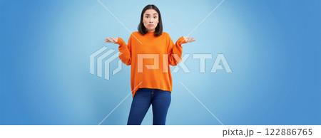 I dont know. Puzzled asian woman shrugs shoulders with hands spread sideways, confused, dont know what to do, clueless and unaware what happened, blue background 122886765