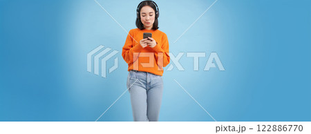 Young woman in headphones and with smartphone listens music, enjoys singing favourite songs in her playlist, blue background 122886770