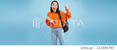 Enthusiastic girl student, celebrating, wearing backpack and holding notebooks, college homework, standing over blue background Enthusiastic girl student, celebrating, wearing backpack and holding notebooks, college homework, standing over blue background 122886793