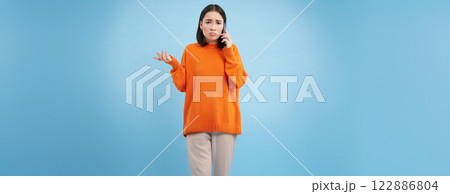 Sad and confused asia girl shrugs, talks on cellphone, answers telephone call with puzzled face, stands over blue background 122886804