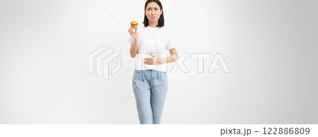 Portrait of asian woman feeling pain in stomach, discomfort after food, holding cupcake and looking unhappy, white background 122886809