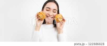 Image of happy korean girl who loves pastry, smiling and eating cupcakes, isolated on white background Image of happy korean girl who loves pastry, smiling and eating cupcakes, isolated on white background 122886828