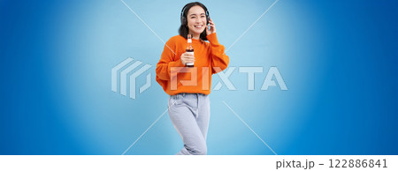 Stylish young female student, girl in headphones drinks from bottle and smiles, stands with beverage against blue background Stylish young female student, girl in headphones drinks from bottle and smiles, stands with beverage against blue background 122886841