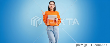 Portrait of korean woman with digital tablet, pointing at upper left corner with confused, frowning face, standing over blue background Portrait of korean woman with digital tablet, pointing at upper left corner with confused, frowning face, standing over blue background 122886843