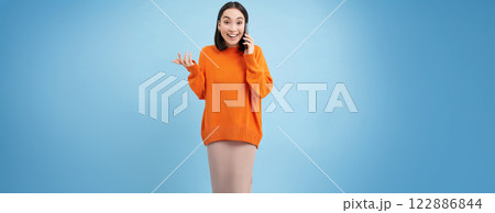 Korean girl in orange sweater talks on mobile phone, speaks on cellphone with happy smiling face, orders delivery, consults with someone on telephone 122886844