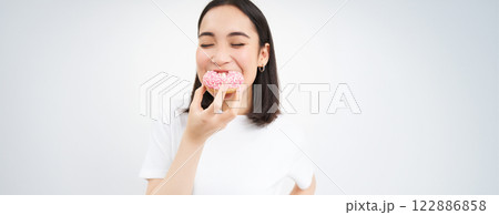 Food and eating out. Young korean woman looks, takes bite of delicious pink glazed doughnut, white background Food and eating out. Young korean woman looks, takes bite of delicious pink glazed doughnut, white background 122886858
