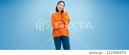 Beautiful smiling asian woman holds mobile phone with happy face. Girl talking on smartphone, listening to voice mail, standing over blue background 122886874