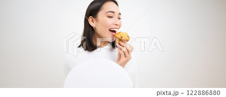 Close up of happy korean woman eating cupcake, delicious dessert, smiling from pleasure, white background Close up of happy korean woman eating cupcake, delicious dessert, smiling from pleasure, white background 122886880