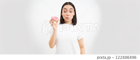 Food and eating out. Young korean woman looks, takes bite of delicious pink glazed doughnut, white background 122886896