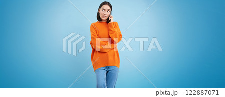 Girl with puzzled face answers phone call, looks confused while talks on cellphone, stands over blue background 122887071