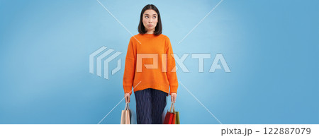 Clueless asian girl looks aside with puzzled face, pouts confused, stands in orange sweatshirt against blue background 122887079