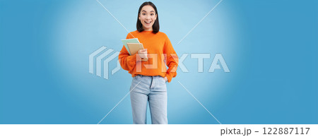 Young asian student with notebooks, standing in orange sweatshirt, looking at camera, posing over blue background 122887117