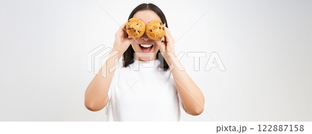 Close up portrait of funny asian girl, holds cupcakes near face, makes happy faces, white studio background Close up portrait of funny asian girl, holds cupcakes near face, makes happy faces, white studio background 122887158