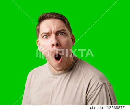 A surprised man stands in front of a bright green screen, expressing shock with his mouth open 122888574