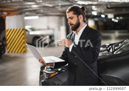 Young caucasian male entrepreneur stands in parking lot engaged in professional video call using laptop. Urban setting emphasizes modern remote work lifestyle. 122899087