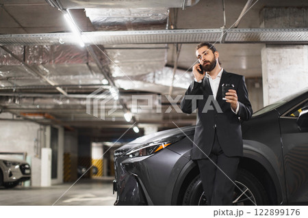 Middle-aged Caucasian male in a suit using smartphone in a parking garage. Professional appearance and modern environment represent business and communication concepts. Middle-aged Caucasian male in a suit using smartphone in a parking garage. Professional appearance and modern environment represent business and communication concepts. 122899176