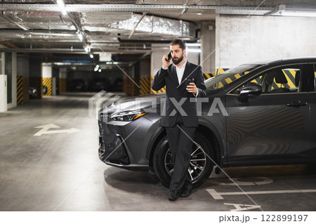 Young adult Caucasian male in business suit talks on smartphone in underground parking beside parked car. Business communication in urban setting. Young adult Caucasian male in business suit talks on smartphone in underground parking beside parked car. Business communication in urban setting. 122899197