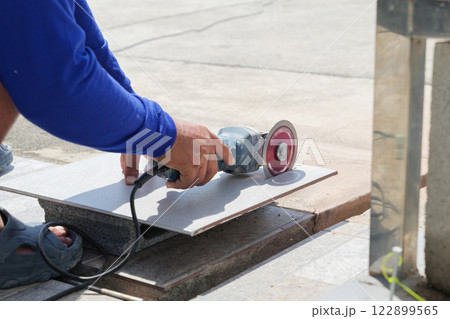 A worker wearing blue clothing operates an angle grinder to cut a gray tile on a piece of stone. The close-up view emphasizes the precision and craftsmanship of tile cutting at a construction site. 122899565