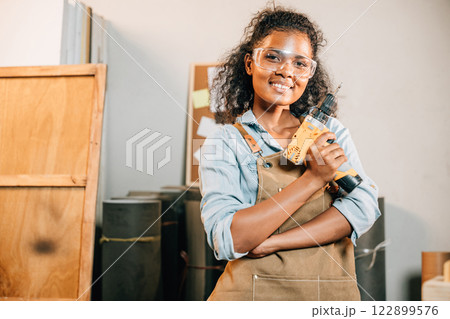 Smiling carpenter woman holding a power drill in a workshop, with a wooden stool and small house models nearby. Surrounded by tools, showcasing her craft and enthusiasm for woodworking projects Smiling carpenter woman holding a power drill in a workshop, with a wooden stool and small house models nearby. Surrounded by tools, showcasing her craft and enthusiasm for woodworking projects 122899576