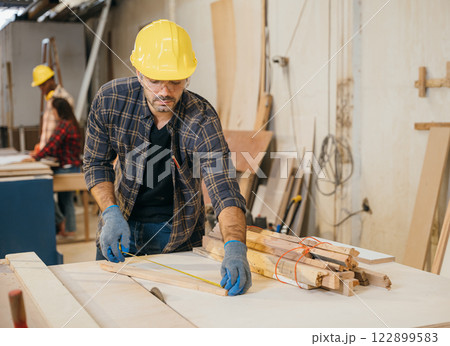 Carpenter wearing a yellow hard hat and safety glasses measuring wood with tape measure. Ideal for woodworking, furniture making, and cabinetry, showcasing precision. Labor day Carpenter wearing a yellow hard hat and safety glasses measuring wood with tape measure. Ideal for woodworking, furniture making, and cabinetry, showcasing precision. Labor day 122899583