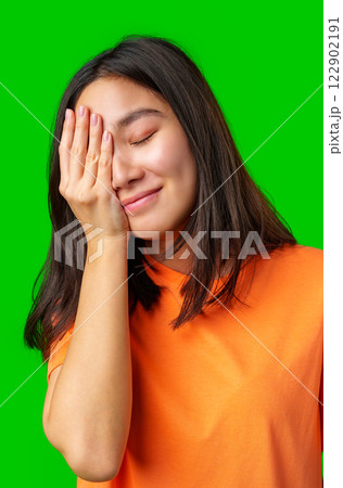 Young woman smiling with one hand on her face against a bright green background during a lighthearted moment 122902191