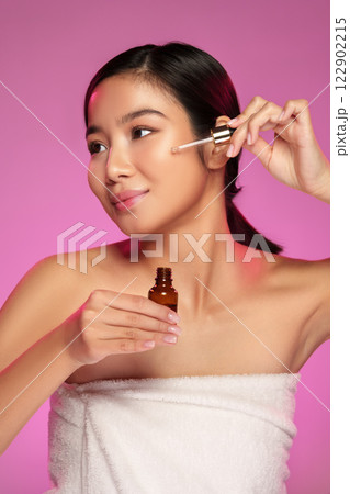 Woman applies serum to her face while smiling against a bright pink background in a beauty routine Woman applies serum to her face while smiling against a bright pink background in a beauty routine 122902215