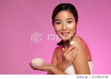 Woman applying body cream on her shoulder with a cheerful expression in a pink backdrop during a skincare routine 122902336