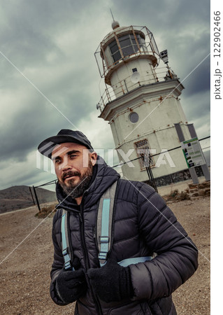 Man stands near an old lighthouse on a cloudy day while wearing a jacket and cap, showcasing the rugged coastal landscape Man stands near an old lighthouse on a cloudy day while wearing a jacket and cap, showcasing the rugged coastal landscape 122902466